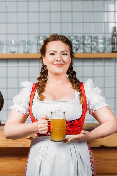 smiling oktoberfest waitress in traditional bavarian dress showing mug of light beer near bar counter
