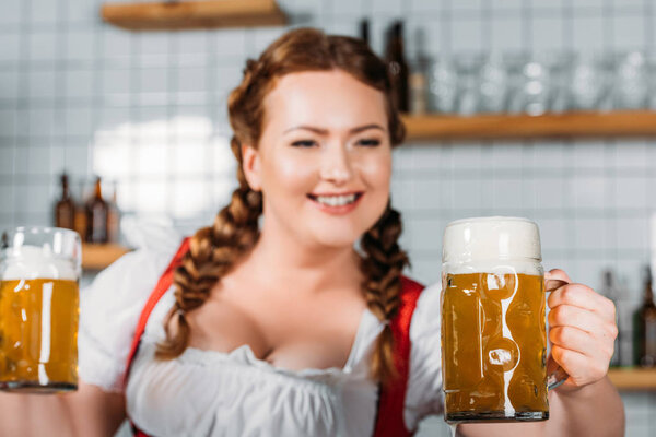 selective focus of oktoberfest waitress in traditional bavarian dress showing mugs of light beer near bar counter