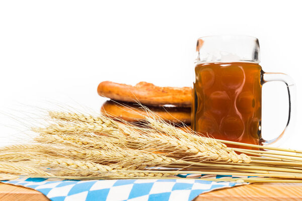close up view of wheat ears, mug of beer and pretzels on table with table cloth on white background 