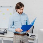 Joven hombre de negocios guapo leyendo documentos en la carpeta en la oficina moderna