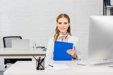 mutlu genç iş kadını işyerinde otururken kameraya bakarak klasörü