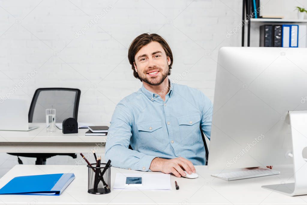 smiling young businessman looking at camera while working with computer at modern office
