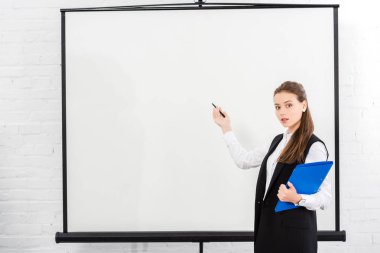 attractive young businesswoman with folder pointing at blank whiteboard at modern office clipart