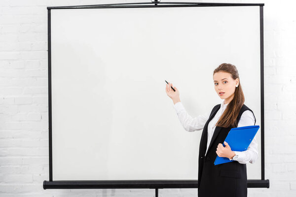 attractive young businesswoman with folder pointing at blank whiteboard at modern office