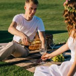 Happy groom pouring wine into glass for young bride on picnic blanket