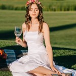 Bela noiva macia em vestido branco e grinalda segurando copo de vinho e sentado em cobertor em piquenique romântico