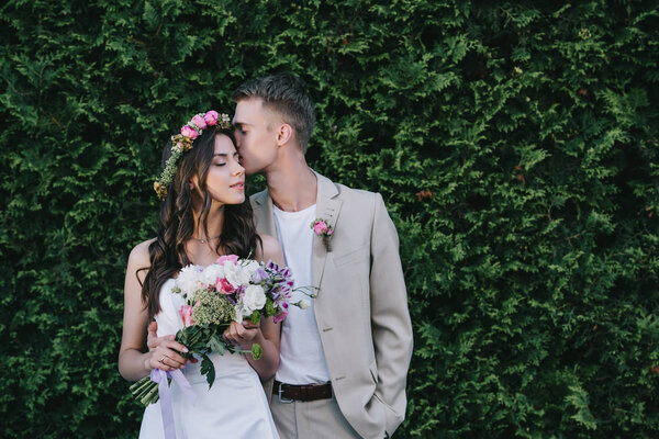 groom kissing and embracing attractive bride in wedding dress with wreath and bouquet