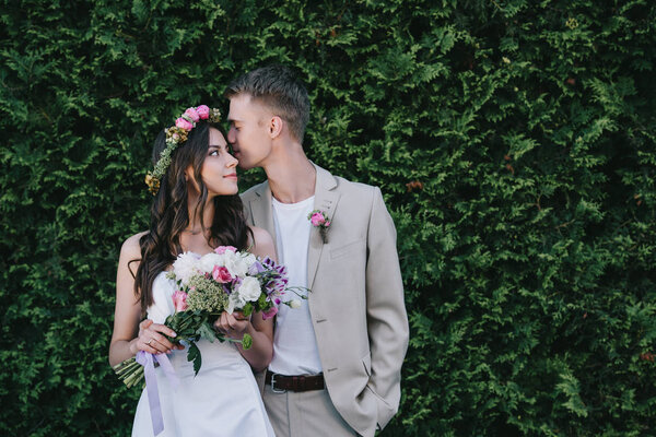 groom kissing and hugging beautiful bride in traditional dress with wreath and wedding bouquet