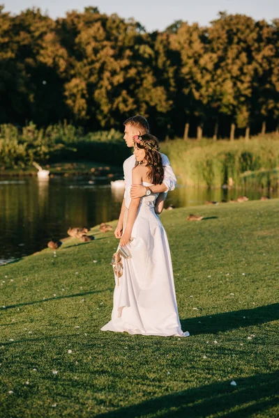 公園の湖の近くに立っている間を受け入れる美しい幸せな若い結婚式のカップル  — 無料ストックフォト