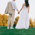 Back view of young wedding couple holding hands and standing on green lawn