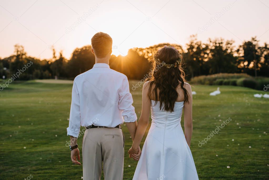 back view of young wedding couple holding hands and standing together in park at sunset 