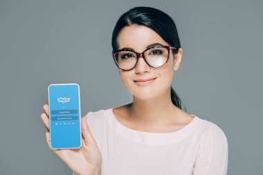 portrait of smiling woman in eyeglasses showing smartphone with skype app on screen isolated on grey clipart