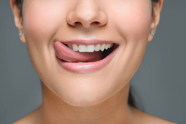 частичный вид улыбающейся женщины с белыми зубами, торчащими языком, изолированный на сером
