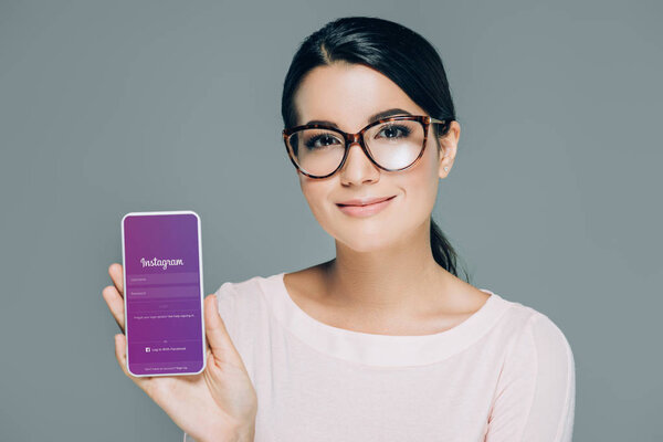 portrait of smiling woman in eyeglasses showing smartphone with instagram app on screen isolated on grey