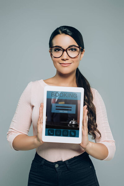 portrait of smiling brunette woman in eyeglasses showing tablet with booking website on screen in hands isolated on grey