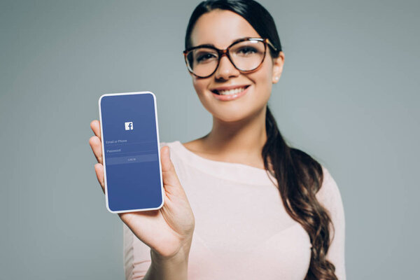 beautiful smiling girl presenting smartphone with facebook app, isolated on grey