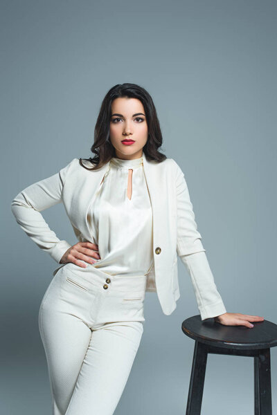 elegant girl in white suit posing at stool, isolated on grey 