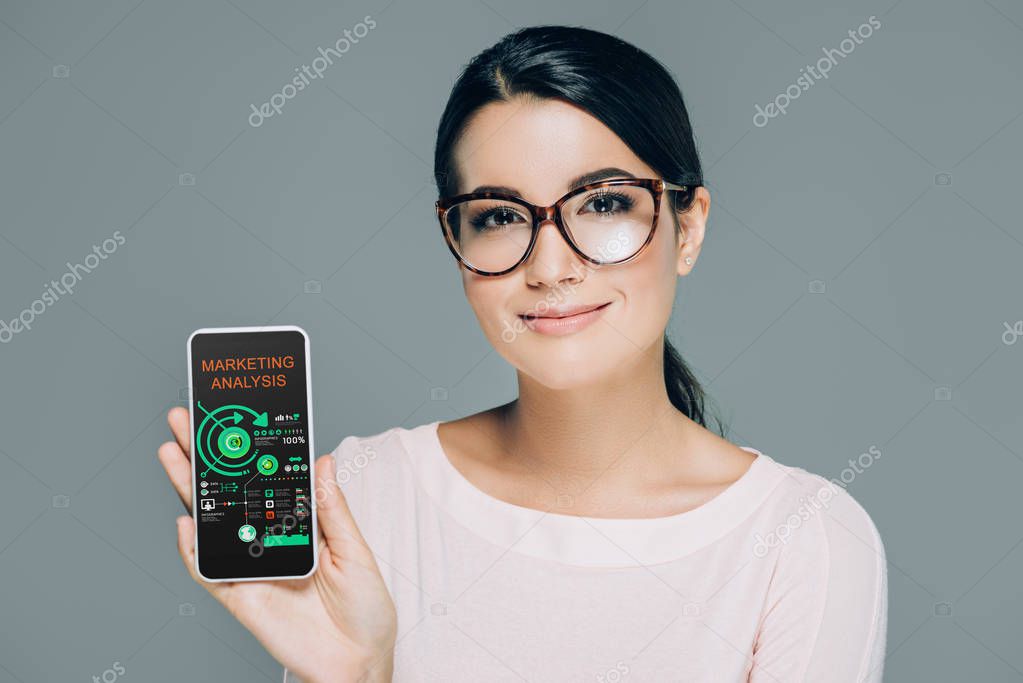 Portrait of smiling woman in eyeglasses showing smartphone with marketing analysis on screen isolated on grey