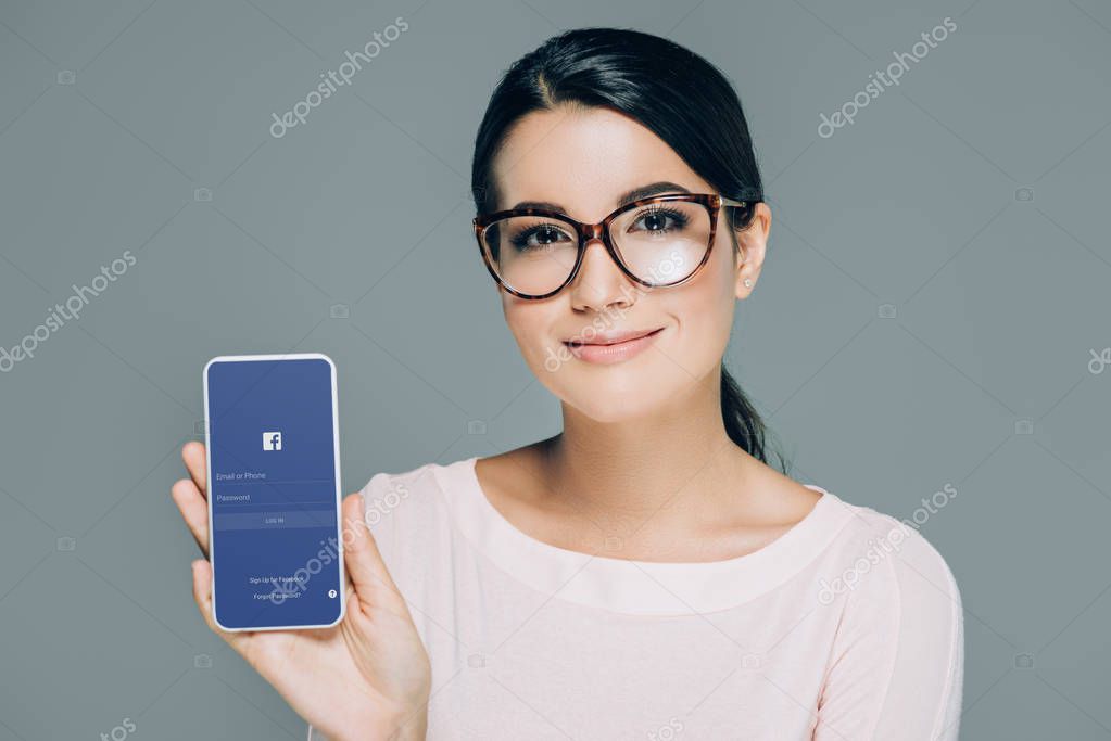 Portrait of smiling woman in eyeglasses showing smartphone with facebook logo on screen isolated on grey