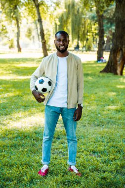 gülümseyen yakışıklı Afrikalı-Amerikalı adam parkta futbol topu ile ayakta