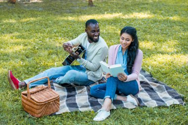 Akustik gitar ve kız okuma kitabı parkta oynarken mutlu Afro-Amerikan erkek