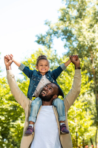 happy african american father holding daughter on shoulders in park 