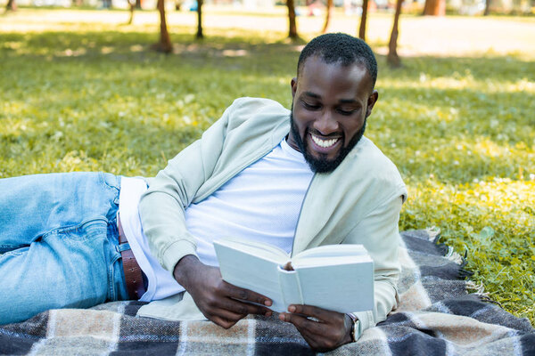 cheerful handsome african american man lying on blanket in park and reading book
