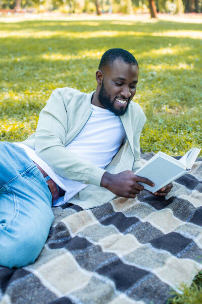 smiling handsome african american man lying on blanket in park and reading book