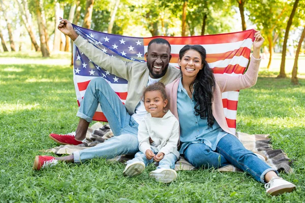 アフリカ系アメリカ人の両親と娘は 公園でのピクニックでアメリカの国旗を保持している笑みを浮かべてください — ストック写真