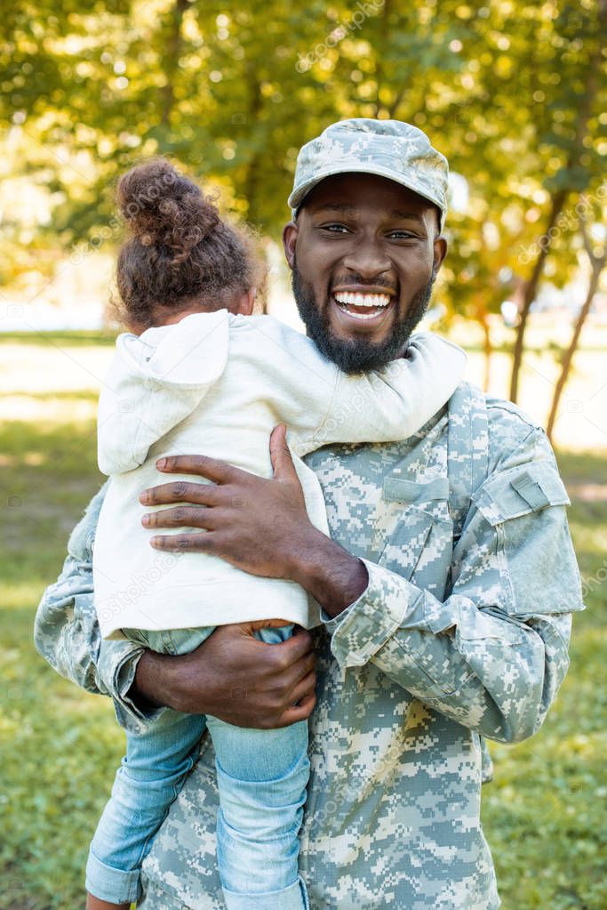 smiling african american soldier in military uniform holding daughter in park