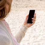 Visão parcial da mulher usando smartphone com tela em branco na praia arenosa