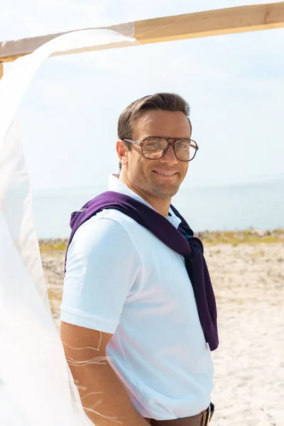 smiling man in eyeglasses standing near decoration with white curtain lace on sandy beach