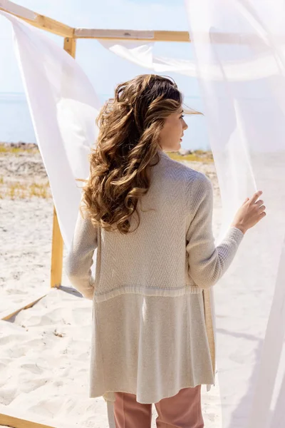 back view of woman standing near wooden decoration with white curtain lace on sandy beach