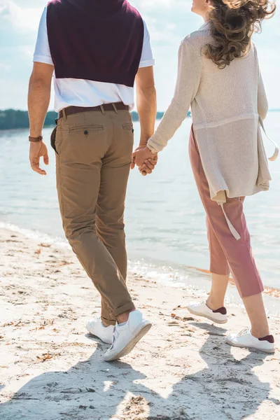砂の川沿いを歩きながら手を繋いでいるロマンチックなカップルの部分的なビュー — ストック写真