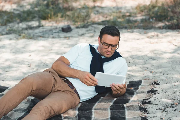 Stilvoller Mann Mit Tablette Während Auf Decke Auf Sandigem Boden — kostenloses Stockfoto