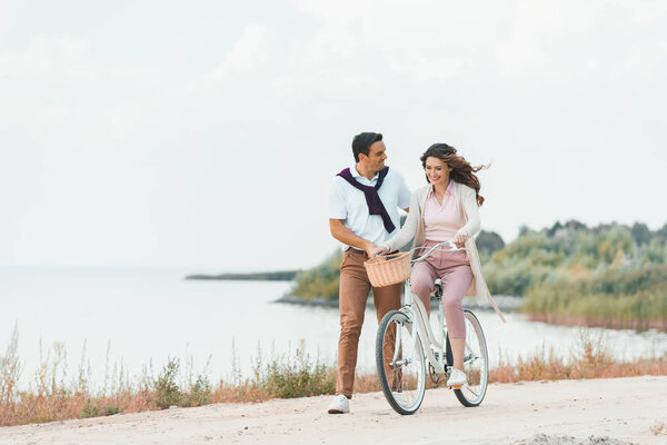 man helping wife ride retro bicycle on sandy riverside 