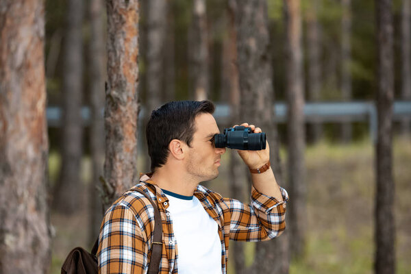 side view of tourist with backpack looking through binoculars in forest