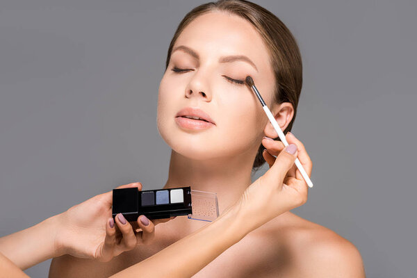 cropped shot of makeup artist applying eyeshadows on models eyelid isolated on grey