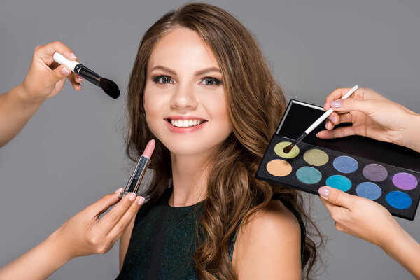 partial view of makeup artists with cosmetics and beautiful smiling woman isolated on grey