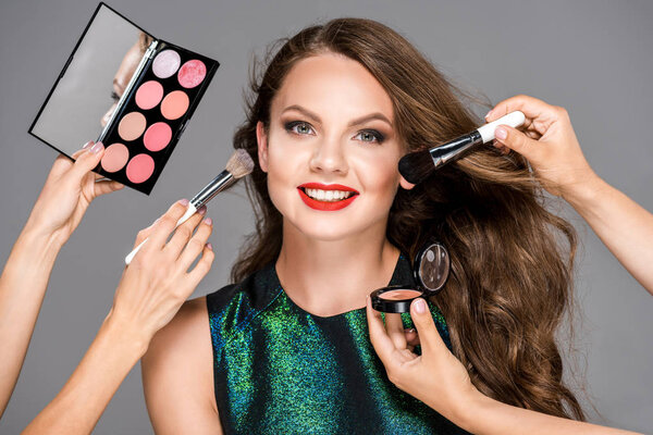 partial view of makeup artists with cosmetics and beautiful smiling woman isolated on grey