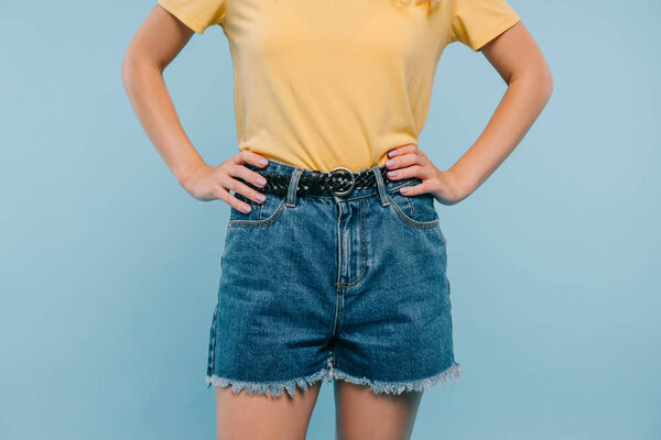 cropped image of girl in shirt and shorts standing with hands akimbo isolated on blue
