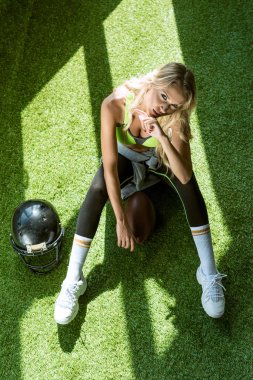 Amerikan futbolu topu ve kask ile yeşil çimenlerin üzerinde oturan uygun genç kadın havai görünümü