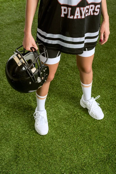 cropped shot of woman in american football uniform holding helmet while standing on field