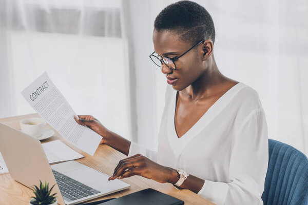 stylish attractive african american businesswoman holding contract and using laptop in office