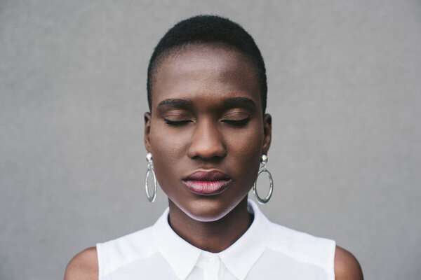 portrait of stylish attractive african american woman standing with closed eyes near grey wall