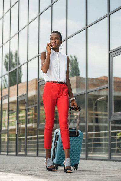 stylish attractive african american businesswoman talking by smartphone and walking with luggage near business center