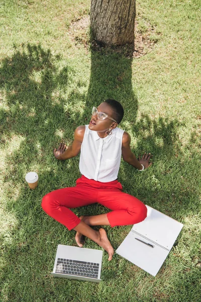 スタイリッシュな魅力的なアフリカ系アメリカ人実業家と見上げると公園の草の上に座ってのハイアングル — ストック写真