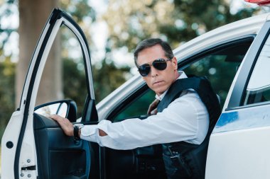 selective focus of mature policeman in bulletproof vest and sunglasses walking out from car  clipart