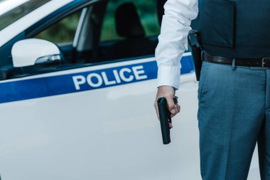 cropped image of policeman in bulletproof vest holding gun near car at street clipart