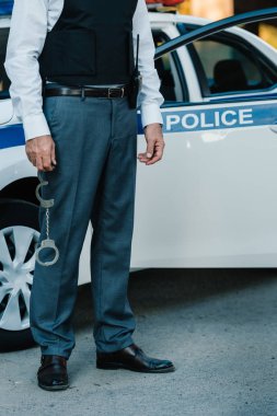 cropped image of male police officer holding handcuffs near car at street clipart
